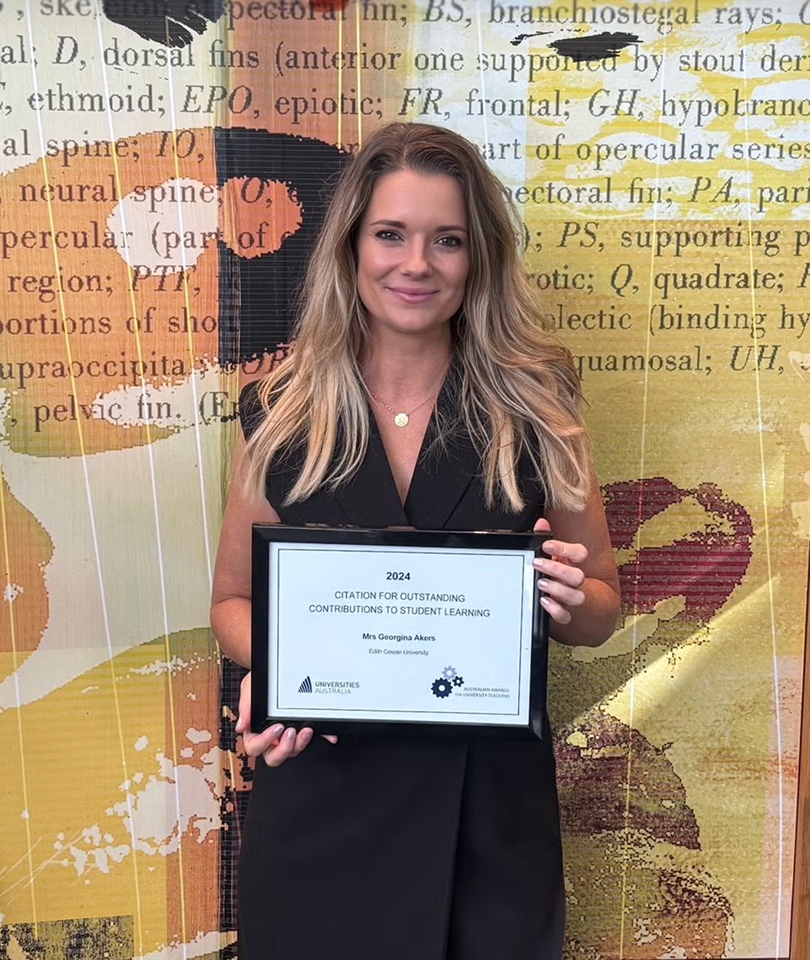 Woman in black dress holding framed 2024 Citation for Outstanding Contributions to Student Learning award certificate in front of decorative patterned wall backdrop