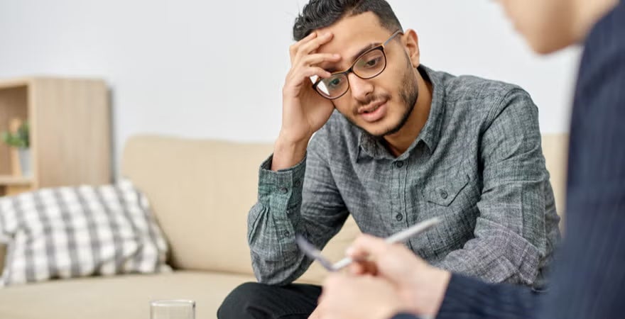 Man in glasses looking worried, sits attentively on a beige couch during a therapy session with a therapist opposite him.