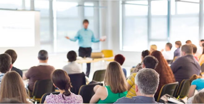 Back view of diverse audience seated and watching male presenter in blue shirt during seminar or workshop in bright modern space