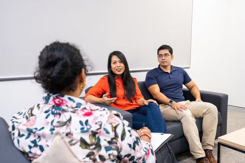 A female counsellor sitting across from a male client in an office, listening attentively and gesturing with her hands as they talk.