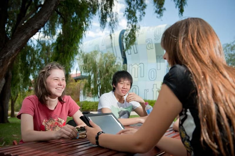 A counsellor sitting with a group of young children outdoors, all smiling and interacting together in a green, sunny setting.