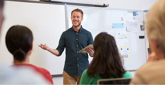 Smiling male instructor in plaid shirt presenting to students in casual classroom training session with whiteboard