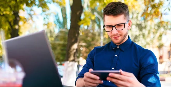 Man in blue shirt wearing glasses using tablet outdoors in natural setting with blurred background