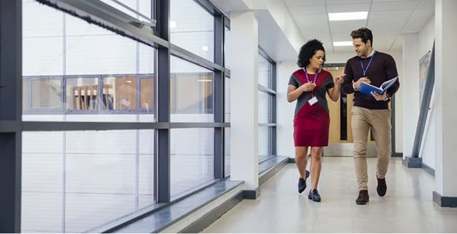 Two diverse professionals walking and discussing while reviewing documents in bright modern office hallway