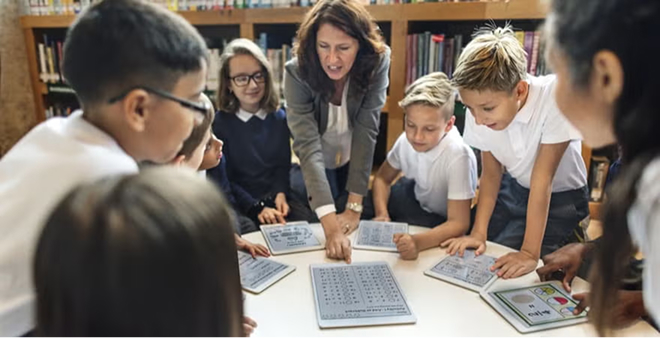 Female teacher in grey vest engaging with diverse group of school children working on tablets and laptops in school library