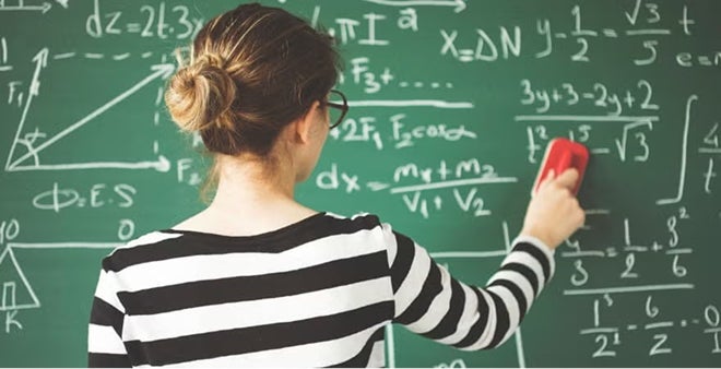Back view of female student with hair in bun wearing striped shirt erasing mathematical equations from green chalkboard