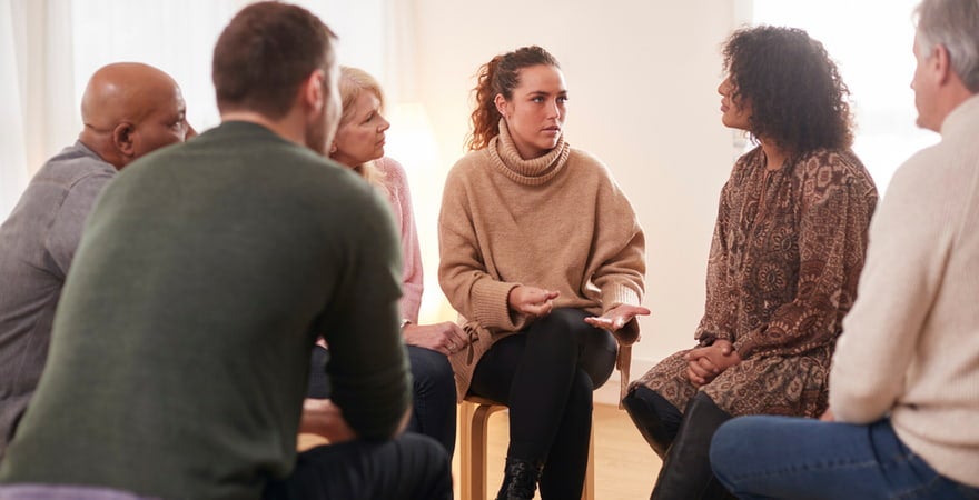 A counselling group sits in a circle, listening intently to a woman in the middle who is talking.