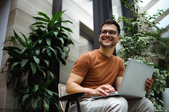 A man sitting on a chair, focused on his laptop, with a neutral background.