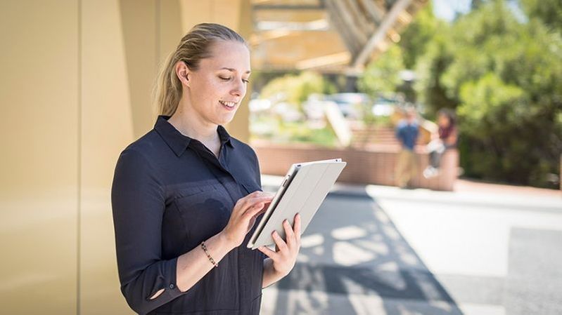 A counselling student is standing outside a building, using her tablet device and smiling. 