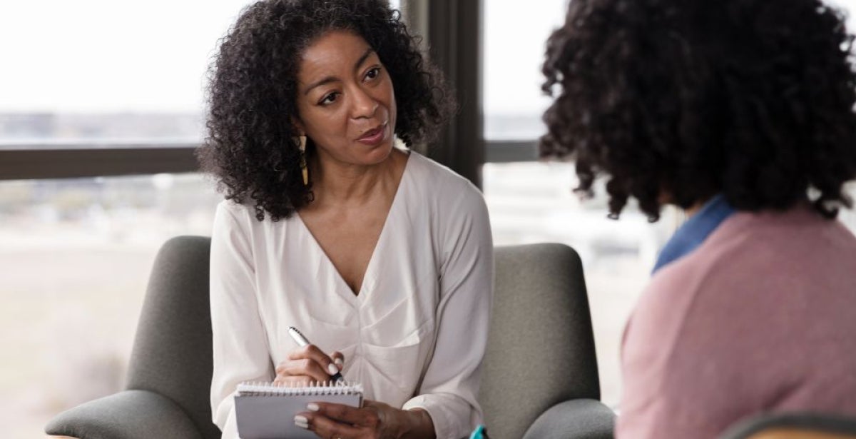 A person in a white top sits in a chair holding a notepad and pen, facing another person whose back is to the camera.