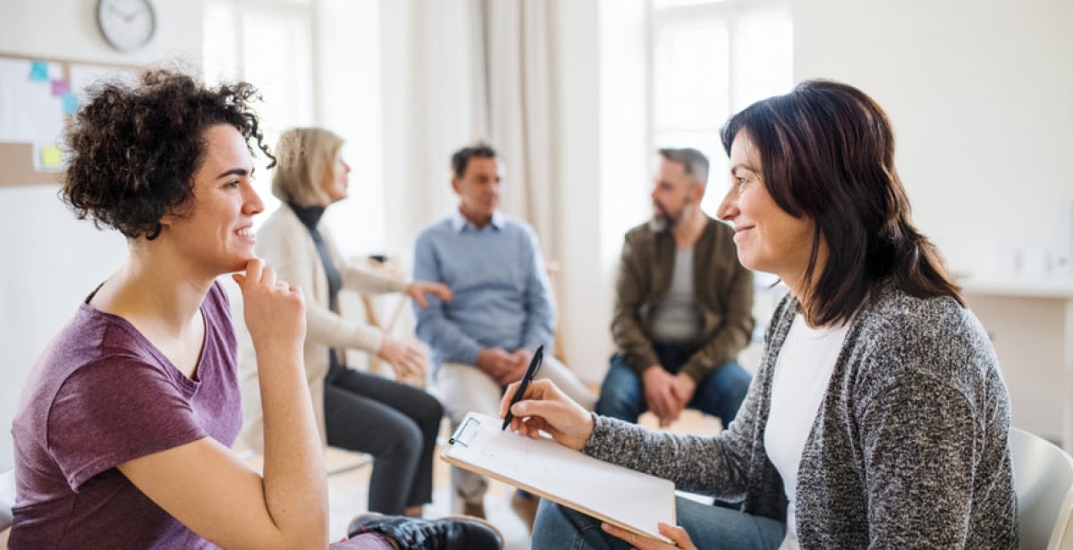 A woman sits in her chair, tlking to a counsellor. They are both smiling and there are people sitting and talking in the background.