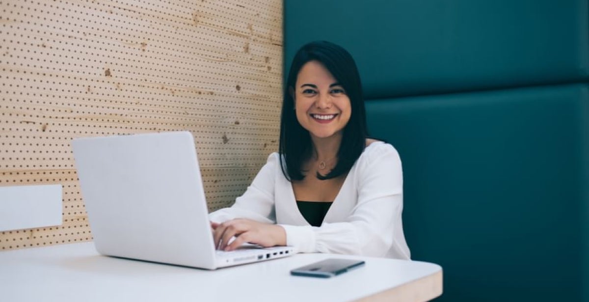 An ECU Online Master of HRM student sits at a table working on her laptop and smiling at the camera.