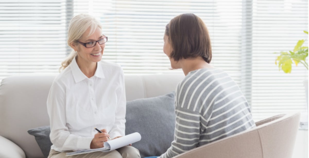 A counsellor is sitting on a couch opposite a client. She is taking notes and smiling.