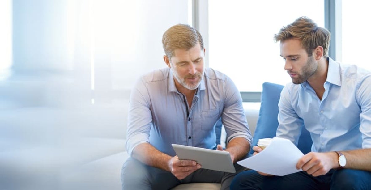 Two men are sitting down together. One is holding a tablet and teaching the other about cyber security. 