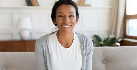 Smiling Black woman with braided hair wearing a white top and gray cardigan, sitting on a beige couch in a cozy living room with plants and a lamp in the background.