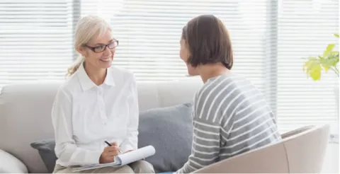 Smiling counselor with glasses and blonde hair in white blouse sitting with client in striped shirt on light grey couch in bright office