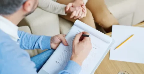 Counsellor taking notes on a clipboard while a client sits nearby with hands clasped.
