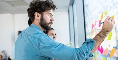 Man in blue shirt writing on colorful sticky notes on glass wall during collaborative workshop session