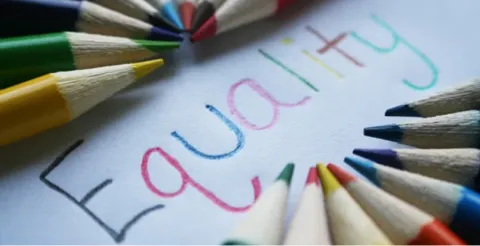 Close-up of equality word written in colorful letters with rainbow of colored pencils arranged around paper on white surface