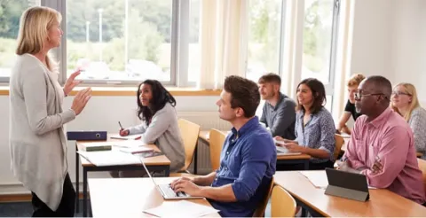 Professional woman instructor in grey cardigan presenting to diverse group of adult students in bright classroom with large windows