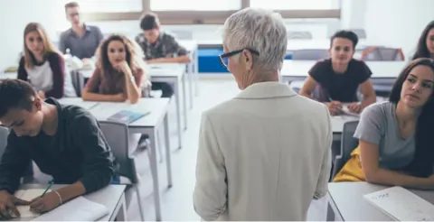 Back view of senior male instructor in beige cardigan teaching diverse adult students in classroom setting