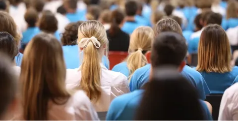 Back view of diverse group of children in blue and white uniforms seated in school assembly or auditorium