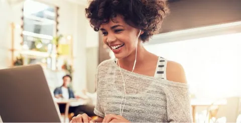 Smiling woman with curly hair wearing earbuds working on laptop in bright modern cafe or office space