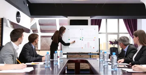 Business professionals in formal attire seated at conference table while woman presents data on whiteboard during corporate meeting