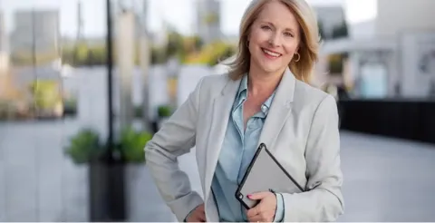 Smiling professional woman in grey blazer holding tablet walking confidently outdoors near modern office building