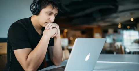 Young man with headphones sitting thoughtfully with hands clasped at laptop in modern cafe or coworking space
