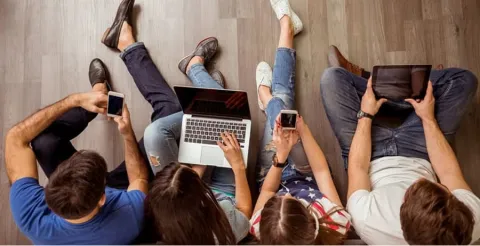 Overhead view of students sitting on wooden floor using laptops, tablets, and smartphones